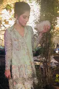 This short frok is decorated with thread, kora, dabka, tilla, sequins and swarovsky. It is embellished with floral thread embroidery. Sleeves are fully embroidered and adorned with cold shoulder design. It is decorated with intricate cutwork on hemline. It is further enhanced with embroidered scalloped border on hemline which adds to the look. It comes with brocade bell shaped pants. It is beautifully coordinated with chiffon dupatta which is sprinkled with sequins all over it.