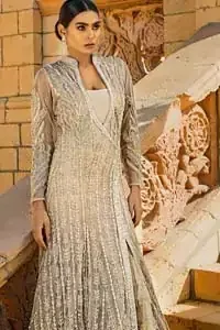 A fusion of traditional and latest fashion making the outfit trendy and unique. This angrakha style floor length maxi is heavily hand crafted with floral embroidery on the bodice, floral booties and vertical lines all over the dress. Back is also enhanced with vertically designed floral booties and swarovski crystals lines all over. The embroidery is done in the shades of silver and dull golden zardozi work. Style it up with frilled lehenga to give it a regal look. This outfit is comprises with fawn net lehenga sprinkled with sequins spray all over.