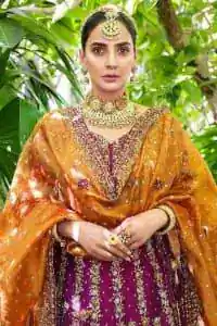 The outfit defines the true essence of allure with its spellbinding magenta hue and intricate details. The blouse is decorated with golden embroidery based on the neckline and sleeves. The sleeves are further enhanced with the embellished handwork. It is Beautifully organized with a magenta lehenga that is again heavily embellished with golden embroidery that is just adding poise to the whole look. Paired along with an orange dupatta decorated with subtly adorned motifs and embellished border that is completing this Mehandi looks wondrously.