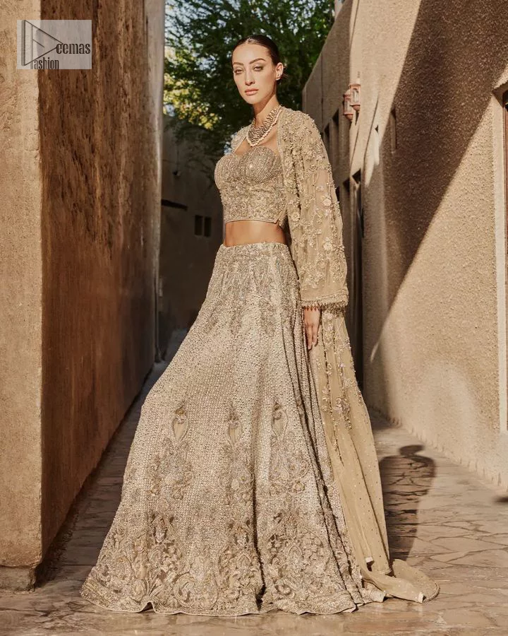 This modern piece is all about extraordinary details and aesthetic vibes! A beige organza blouse is heavily embellished with dabka, naqshi, tilla, Resham, dabka, sequin and Kundan. This gown blouse dress has Mughal-inspired motifs, geometrical patterns and floral motifs. The blouse has a sweetheart neckline or a more upscale appearance and feel. Further, the gown has full sleeves adorned with floral patterns. It is supported by a flared lehenga with an embellished border for additional comfort.