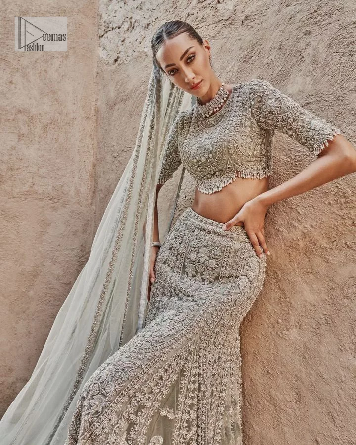 This fusion of floral embroidery is total fashion goodness for a walima outfit! A stunning teal grey organza choli embellished with intricate detailing finished with a scalloped border. The beautiful blouse is decorated with silver embellishment which is further prominent with tilla,daka, kora, Kundan and the real magic of Zardozi.  It is paired with a heavily embellished back-train lehenga with botanical motifs and geometric patterns. Its dupatta is made from a net. The dupatta is a wide embellished border that added divine charm to the entire ensemble.